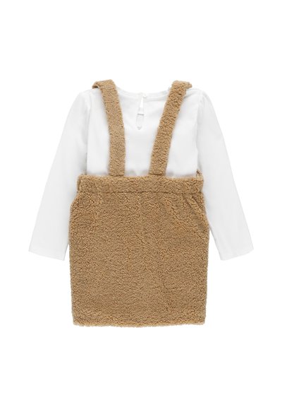 Pinafore in finta pelliccia color tan con parte anteriore testurizzata, bretelle e una camicia a maniche lunghe bianca sotto. Presenta una chiusura con bottoni sul retro.