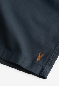Tissu bleu foncé avec une petite tête de cerf brodée en marron près de l'ourlet, montrant des coutures détaillées et une texture riche.