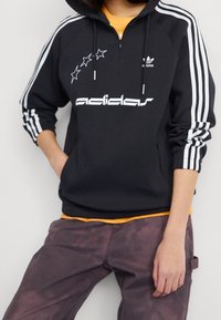 Black hooded sweatshirt made of fabric, featuring white Adidas logo and stars, with three white stripes on sleeves and a zippered front.