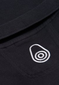 Tela negra con un tejido texturizado que presenta un logo de aguacate bordado en blanco con círculos concéntricos, ubicado cerca del cuello.