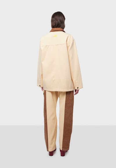 Ljust gul oversized jacka med brun krage, kombinerad med beige och brun panelbyxor. Noterbara knappstängningar och textlogotyp på baksidan.