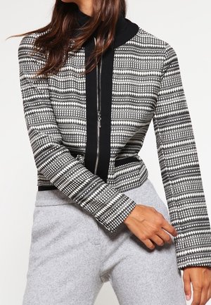 Woman wearing a black and white patterned zip-up jacket with black trim and light gray pants, posing with left arm across body.