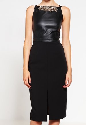Mujer con un vestido negro sin mangas con la parte superior de aspecto cuero y adorno con joyas en el escote, y una falda hasta la rodilla con una abertura frontal.