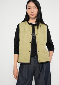 Prošívaná vesta ve světle olivově zelené barvě s kulatým výstřihem, zapínáním na druky a bočními kapsami, spárovaná s černým tričkem s dlouhým rukávem a tmavými kalhotami.