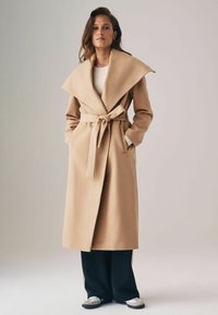 Beige trench coat széles gallérral, övvel a deréknál és oldalsó zsebekkel, sötét nadrággal és hétköznapi lábbelivel párosítva. Simaszerkezet.