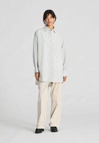 Lange mouw blouse in lichtgrijs met smalle verticale strepen, gecombineerd met beige wijde broekspijpen en zwarte schoenen.