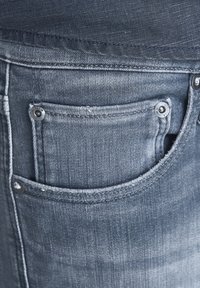 Mörkblå, strukturerade jeans i denim med en synlig bakficka och metallnitar. Tyget ser slitet ut runt fickans kanter, vilket betonar hållbarheten.