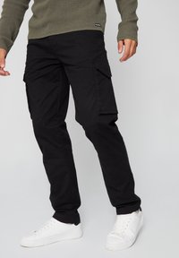 Zwarte cargobroek van duurzaam materiaal, met meerdere zijzakken, gedragen met witte sneakers. De pasvorm is relaxed met een rechte pijp.