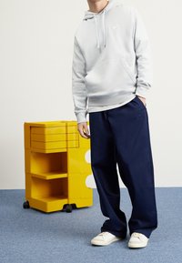 Person i vit Nike-huvtröja, marinblå byxor och vita sneakers står bredvid en gul rullande förvaringsenhet på blå mattlagd golv.