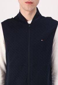 Granatowa kamizelka pikowana z wysokim kołnierzem i zamek błyskawiczny z przodu, z niewielkim czerwono-białym logo po lewej stronie klatki piersiowej. Miękka tekstura.