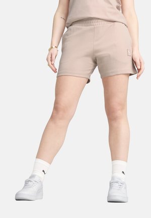 Pantalones cortos deportivos beige con cintura elástica, bolsillo lateral y logotipo blanco, combinados con zapatillas blancas y calcetines blancos acanalados.