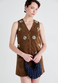 Brown suede sleeveless dress with floral embroidery and a V-neck, paired with a dark blue, textured woven handbag.