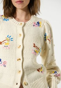 Cardigan en tricot crème avec broderie florale colorée, encolure ronde, fermetures à boutons et motifs texturés sur les manches et le corps.