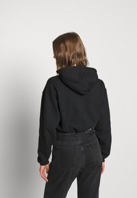 Donna con capelli castani di lunghezza media, che indossa un felpa nera e jeans scuri a vita alta, in piedi e rivolta verso uno sfondo grigio chiaro uniforme.