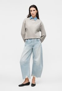 Mujer con cabello largo y oscuro lleva una sudadera gris sobre una camisa azul con cuello, vaqueros cortos de pierna ancha desgastados y zapatos planos negros.