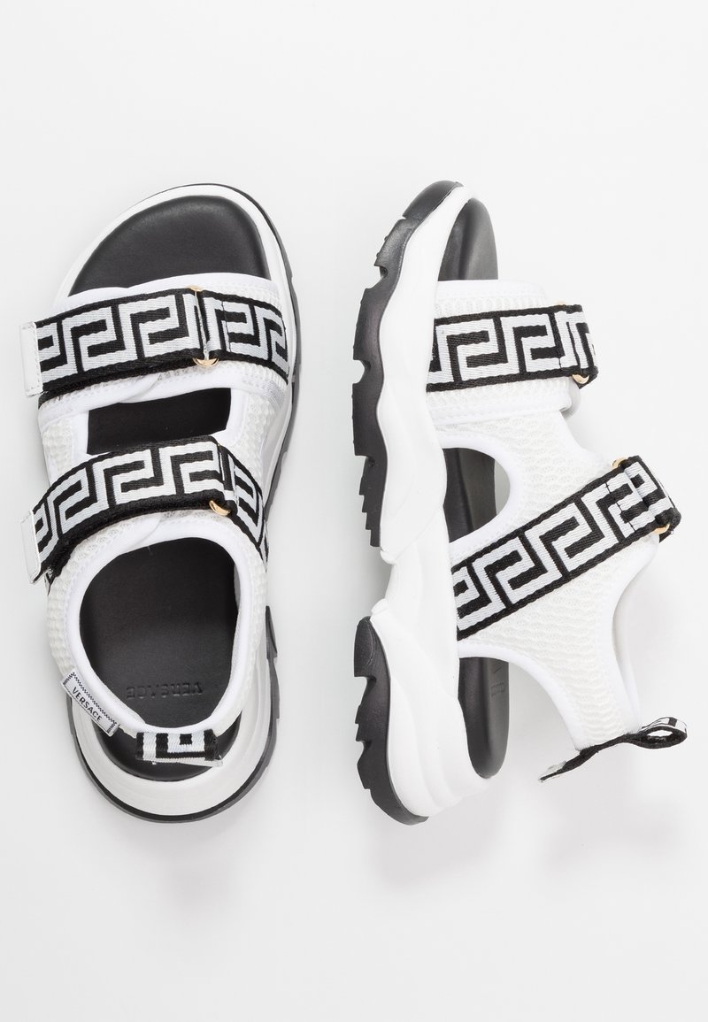 Sandales blanches et noires avec des semelles épaisses, deux bandes velcro avec un motif géométrique grec noir.