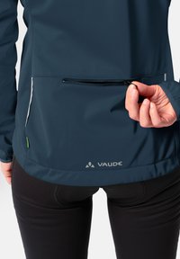 Giacca softshell blu navy con tasca con zip sulla schiena. Presenta una texture liscia, un orlo arrotondato e dettagli riflettenti. Logo Vaude in evidenza.