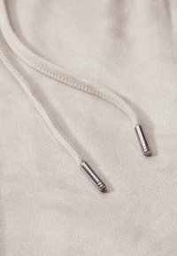 Lys beige hoodie-stof med glat tekstur; har snore med metalagtige ender. Nærbillede fremhæver syningsdetaljer.