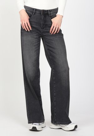 Person iført høj-taljede, vidde, falmede sorte jeans, hvid langærmet top og hvide sneakers, stående med hænderne hvilende nær taljen.