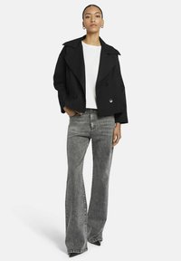 Vrouw staat in een zwarte cropped dubbel-gebreide jas, witte top, grijze hoog-waist flared jeans en zwarte puntige schoenen.