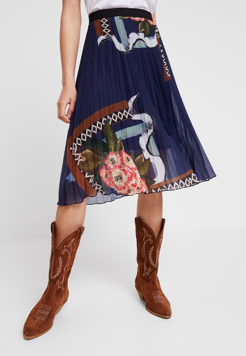 Gonna plissettata blu navy con motivi floreali e geometrici, dotata di una fascia elastica nera in vita. Abbinata a stivali cowboy in suede marrone.