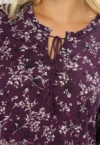 Luonnonvalkoisessa kukallisessa paidassa on avainaukko kaula-aukko ja nauhakiinnitys. Siinä on valkoisia ja vihreitä kukkakuoseja, kevyt kangas ja pehmeä rakenne.