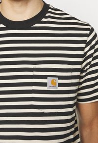 T-shirt à rayures noires et blanches avec un col rond, comportant une poche poitrine et un patch logo Carhartt rectangulaire. Matière en coton.
