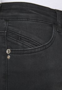 Black denim jeans feature a small front pocket with two silver metal studs and contrasting stitching details along the waistband and seams.