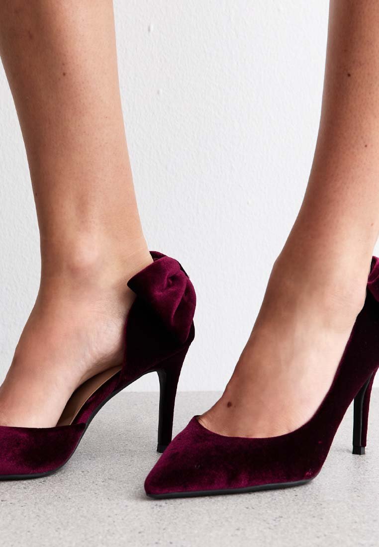 Burgundy sammet högklackade skor med spetsig tå och öppen baksida, med en rosettaccent vid vristen. Svart stilettklack. Slät yta.