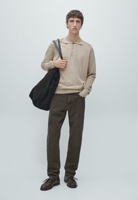 Polo beige lavorata a maglia con tre bottoni, pantaloni marrone scuro a gamba dritta e scarpe di pelle marrone. Una borsa a tracolla in camoscio scuro è appesa a una spalla.