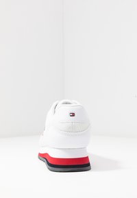 Baskets blanches avec semelle à rayures rouges et noires, détails en mesh et petit logo rectangulaire à l'arrière, vues de dos sur fond blanc.