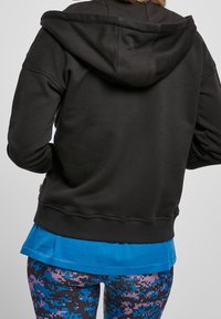 Persona con un hoodie nero sopra una maglietta blu e leggings a motivo pixel in blu, viola e nero, vista di spalle.