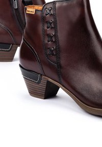 Botas de tobillo de cuero marrón con tacón apilado, costuras decorativas en negro, cremallera lateral y etiqueta de la marca Pikolinos.