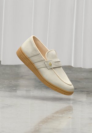 Beige mokkanahkainen Adidas-mokkasiini purukumipohjalla, kolmella sivuraitaprofiililla ja kultaisella logolla remmillä, leijumassa kiillotetun lattian yläpuolella.