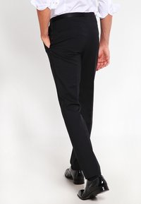 Zwarte pantalon van een gladde stof, met een getailleerde pasvorm, platte voorkant en standaardzakken, gecombineerd met glanzende zwarte schoenen.