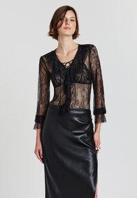 Blusa in pizzo nero con scollatura a V e bordi arricciati, dotata di maniche lunghe e fronte annodata. Abbinata a una gonna in pelle nera elegante.