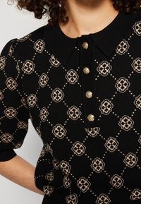 Top en maille noir avec motif floral beige en losange, manches courtes bouffantes, col pointu et quatre boutons décoratifs dorés sur la patte de boutonnage avant.