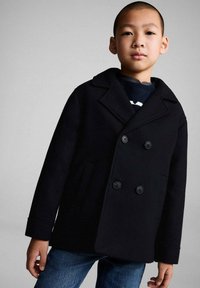 Svart dubbelknäppt kappa i ull, med stora knappar, slagkrage och sidofickor, som bärs tillsammans med jeans.