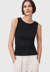 Top nero senza maniche in cotone elastico con scollatura tonda, caratterizzato da dettagli arricciati in vita. Indossato con pantaloni di colore chiaro.