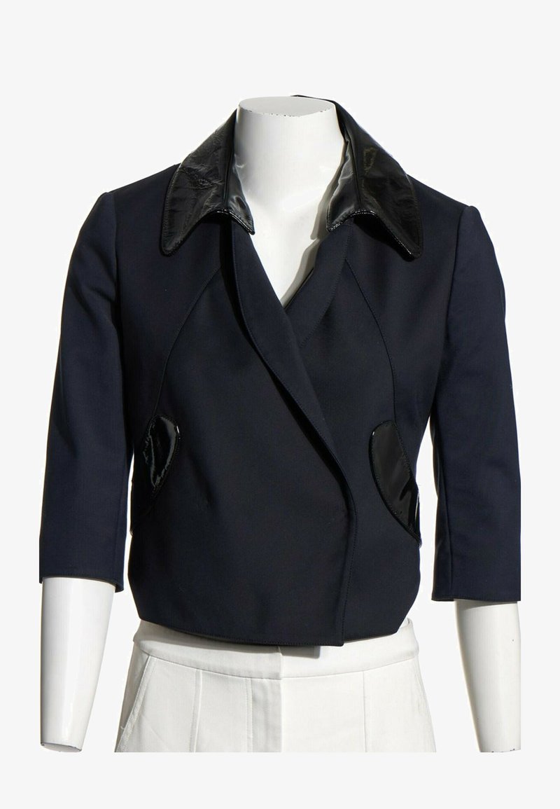 Veste noire courte avec manches trois-quarts, col noir brillant et détails de poche sur un torse de mannequin blanc.
