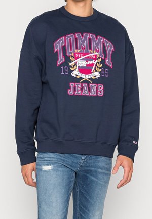Osoba ubrana w granatową bluzę z dużym napisem "Tommy Jeans" i logo herbu, w połączeniu z jasnoniebieskimi podniszczonymi jeansami.