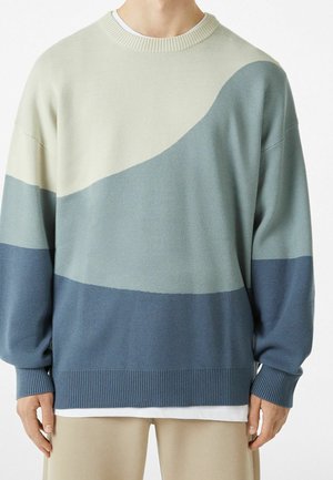 Personne portant un pull ample à manches longues avec des blocs de couleur courbes en crème, bleu clair et bleu foncé, associé à un pantalon beige.
