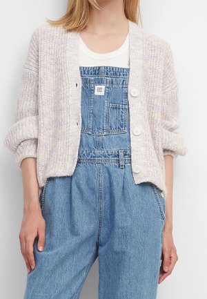 Person iført en hvid strikket cardigan med op-rullede ærmer over en hvid skjorte og blå denim overalls med en forlommelomme.