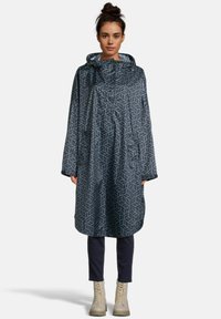 Vrouw in een donkergepatenteerde regenponcho met zakken en capuchon, marineblauwe broek en beige veterschoenen, staande met het gezicht naar voren op een witte achtergrond.