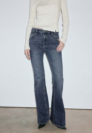 Donna con jeans svasati blu scuro a vita alta e maglia a coste crema a maniche lunghe, con una mano in tasca e scarpe nere appuntite.