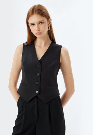 Gilet noir sans manches avec une silhouette ajustée, décolleté en V, cinq boutons sur le devant et deux poches latérales. Tissu lisse avec une finition mate.