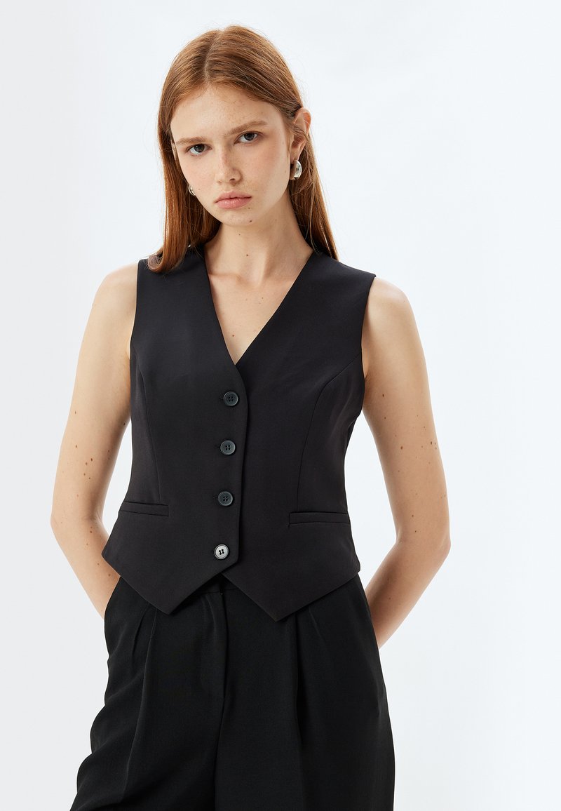 Schwarze, ärmellose Weste mit einer taillierten Silhouette, V-Ausschnitt, fünf Knöpfen vorne und zwei seitlichen Taschen. Glattes Material mit matter Oberfläche.