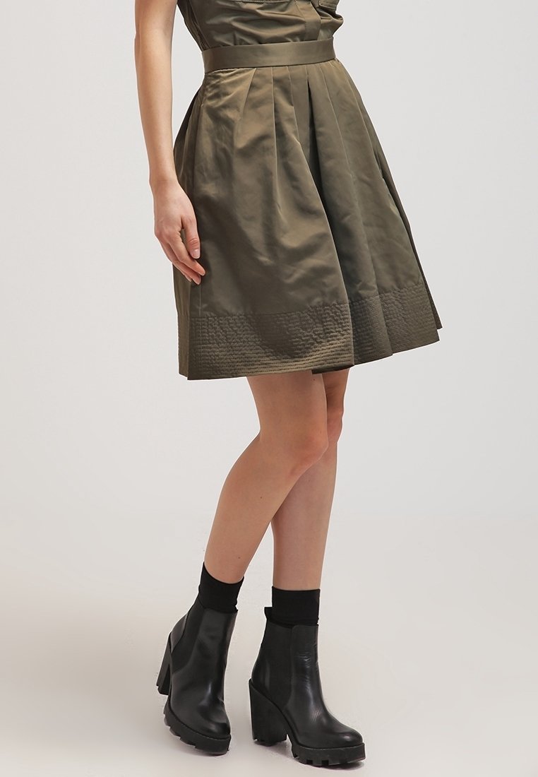 Oliivinvihreä, polvipituinen hame, jossa on laskostettu muotoilu, teksturoitu helma ja siihen kuuluva vyötärövyö. Yhdistetty mustien nilkkureiden ja sukkien kanssa.