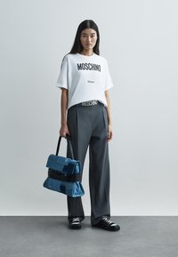 White cotton t-shirt with "Moschino Milano" text, paired with gray pleated trousers. Carrying a blue denim bag with black hardware. Black sneakers.