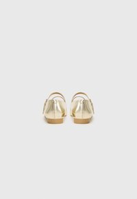 Chaussures plates en métal doré avec une finition lisse, vue arrière montrant une forme arrondie, une sangle en cuir souple, et un petit talon en caoutchouc tan.